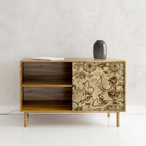 Puede incluir: Un mueble de madera con una puerta corredera con un diseño de mapa antiguo. El mueble tiene dos estantes y se apoya en cuatro patas delgadas. Un jarrón gris oscuro y un libro blanco están encima. El diseño del mapa es en tonos marrones y beige.