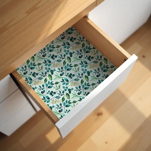 May include: A wooden drawer with a white front is open, revealing a patterned liner. The liner features a repeating design of green and teal leaves and berries on a white background. The drawer is part of a light-colored wooden desk.