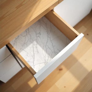 May include: Open wooden drawer with a white marble-patterned liner. The drawer is part of a light-colored wooden desk or cabinet. The drawer front is white, contrasting with the wood grain of the desk and drawer interior.