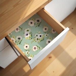 May include: A drawer with a floral patterned liner. The liner features white anemone flowers with black and pink centers, green leaves, and stems on a light green background. The drawer is open, revealing the floral design.