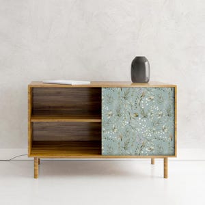 Puede incluir: Gabinete de madera con un diseño floral en la puerta corredera. El gabinete tiene dos estantes y cuatro patas. Un jarrón gris oscuro y un libro blanco están encima. La puerta tiene un fondo azul claro.