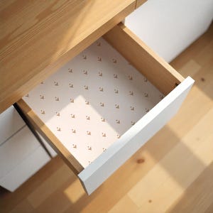 May include: Open wooden drawer with a white front. The drawer interior features a patterned liner with small, brown, stylized letter "L" shapes on a cream background. The drawer is part of a larger wooden desk.
