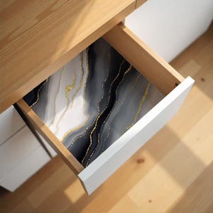 May include: An open white drawer with a decorative liner featuring a marble design. The liner has a pattern of flowing black, gray, and white with gold accents. The drawer is set in a light wood cabinet.