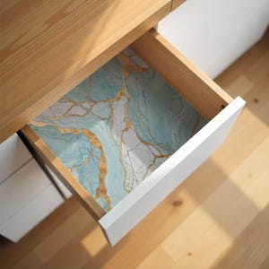 May include: Open drawer with a marble-patterned liner. The liner features a blue, white, and gold design. The drawer is part of a wooden desk with white drawer fronts.