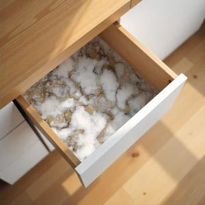 May include: A drawer with a white front and a wooden interior, revealing a marble-patterned liner. The liner features a white base with gold veining, creating a luxurious and decorative effect. The drawer is partially open, showcasing the design.