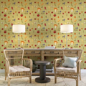 May include: A cozy living room scene featuring two rattan armchairs with cream-colored cushions, a wooden console table, and two lamps. The wallpaper has a repeating pattern of mushrooms, apples, bees, and flowers in red, orange, and blue.