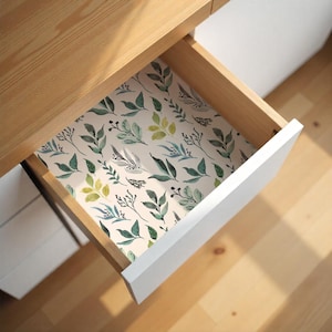May include: An open drawer with a floral patterned liner. The liner features watercolor-style green and blue leaves and sprigs on a cream background. The drawer is white with a wooden frame.