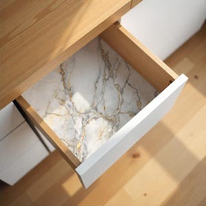 May include: Open wooden drawer with a white front, revealing a marble-patterned liner. The marble design features a mix of white, gray, and gold veining. The drawer is part of a light-colored wooden desk or cabinet.