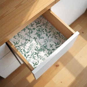 Op de afbeelding: Een open houten lade met een witte voorkant, met een bloemmotief voering. De voering heeft een herhalend ontwerp van groene bladeren en wijnstokken op een witte achtergrond. De lade maakt deel uit van een licht houten bureau.