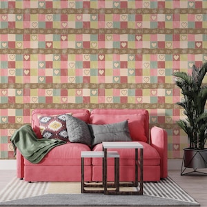 May include: A living room with a pink sofa, cushions, and a green throw. The wall has a patterned wallpaper with squares, hearts, and floral designs in pastel shades. Two small white tables with dark brown legs are in front of the sofa.