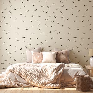 May include: A bedroom scene featuring a bed with a beige knitted throw and pillows. The wall behind the bed is decorated with numerous black bird silhouettes. A woven basket and a lamp are also visible.