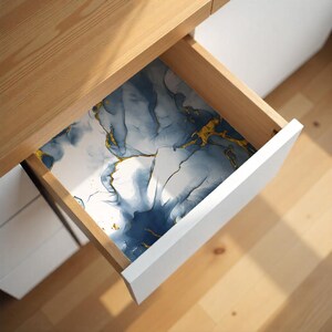 May include: Open drawer with a decorative liner. The liner features a blue and gold abstract design on a white background. The drawer is inside a wooden cabinet with a white drawer front.