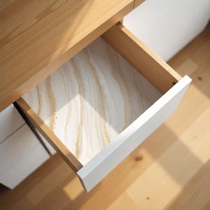 May include: An open wooden drawer with a white front, revealing a decorative liner. The liner features a white background with flowing, gold-colored, glitter-like streaks. The drawer is part of a light-colored wooden desk.