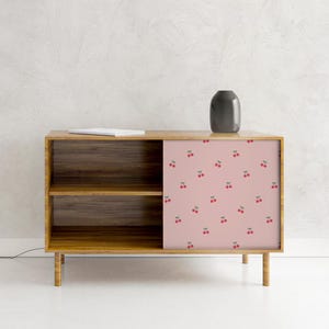 May include: A wooden cabinet with a light brown finish and a sliding door. The door features a pink background with a pattern of red cherries. A dark gray vase sits on top of the cabinet.
