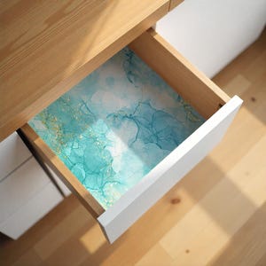 May include: An open wooden drawer with a white front, revealing a decorative liner. The liner features a watercolor design with shades of teal, blue, and gold. The drawer is part of a light-colored wooden desk or cabinet.