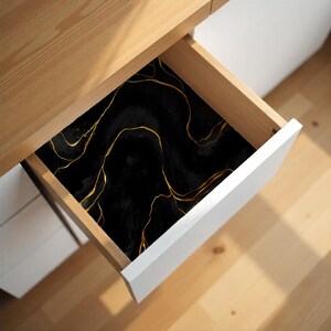 May include: A drawer liner with a black marble design and gold veining. The liner is inside a wooden drawer with a white front. The design has a flowing, abstract pattern.