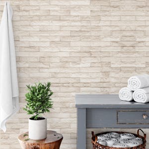 May include: A bathroom scene featuring a light beige brick wall, a small green plant in a white pot, and a wooden stool. A gray table holds rolled white towels. A white towel hangs on the left.