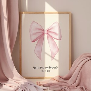 Pode incluir: Uma impressão emoldurada com uma ilustração em aquarela de um laço rosa. A obra de arte inclui o texto "you are so loved. John 3:16" em uma escrita delicada. A moldura é de cor de madeira clara.