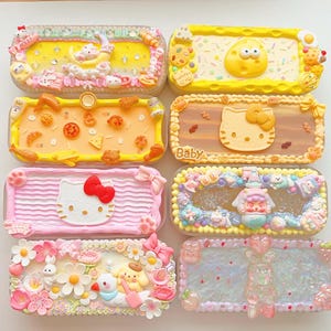 May include: Eight rectangular cases decorated with various miniature charms, including Hello Kitty figures, food items, flowers, and bows. The cases feature textured borders, colourful resin surfaces, and small plastic embellishments in yellow, pink, and blue tones.