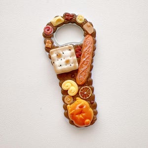 Pu&ograve; includere: Un apribottiglie in metallo decorato con miniature di cibo, tra cui un cracker quadrato, una baguette, una fetta di agrume, un pasticcino e una piccola figura animale arancione, su una base marrone che ricorda una glassa al cioccolato.