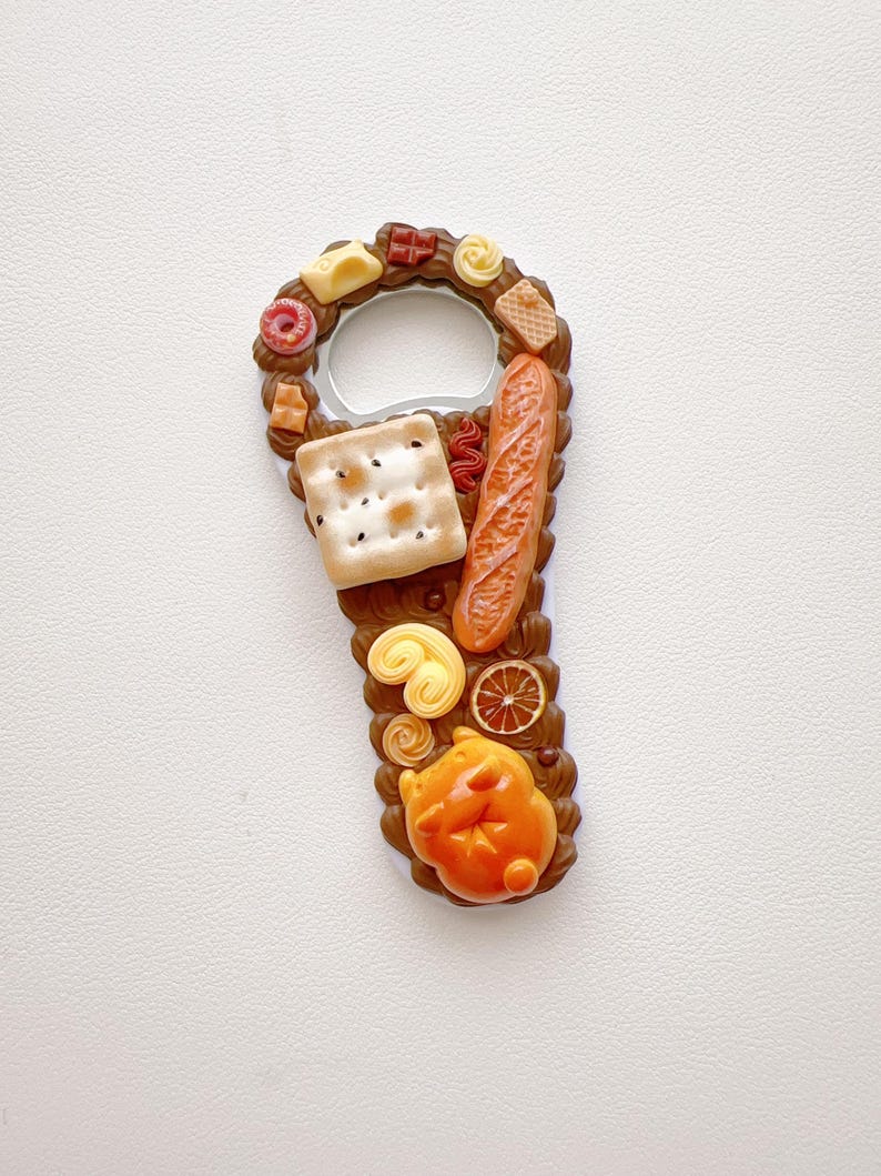 Pu&ograve; includere: Un apribottiglie in metallo decorato con miniature di cibo, tra cui un cracker quadrato, una baguette, una fetta di agrume, un pasticcino e una piccola figura animale arancione, su una base marrone che ricorda una glassa al cioccolato.