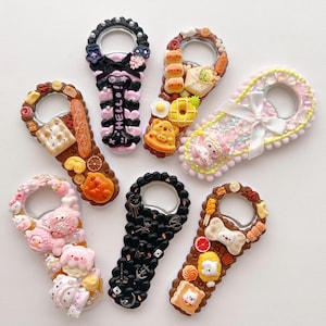 Pu&ograve; includere: Una collezione di sette apribottiglie decorativi con vari ciondoli in miniatura raffiguranti cibo, animali e personaggi. I design includono temi come prodotti da forno, creature rosa, motivi gotici neri e accenti pastello con fiocchi.
