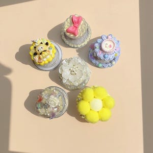 May include: An assortment of decorative phone grips in various designs. The grips feature a yellow pom-pom flower, a white flower with pearl accents, and miniature cakes with bows and animal figures. The grips are set against a neutral background.