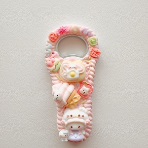 Pu&ograve; includere: Un apribottiglie rosa decorato con ciondoli in resina in miniatura, tra cui una figura di personaggio bianco, un cono gelato, una fetta di pane tostato con un uovo al tegamino e vari vortici colorati e piccole forme di caramelle sui bordi.
