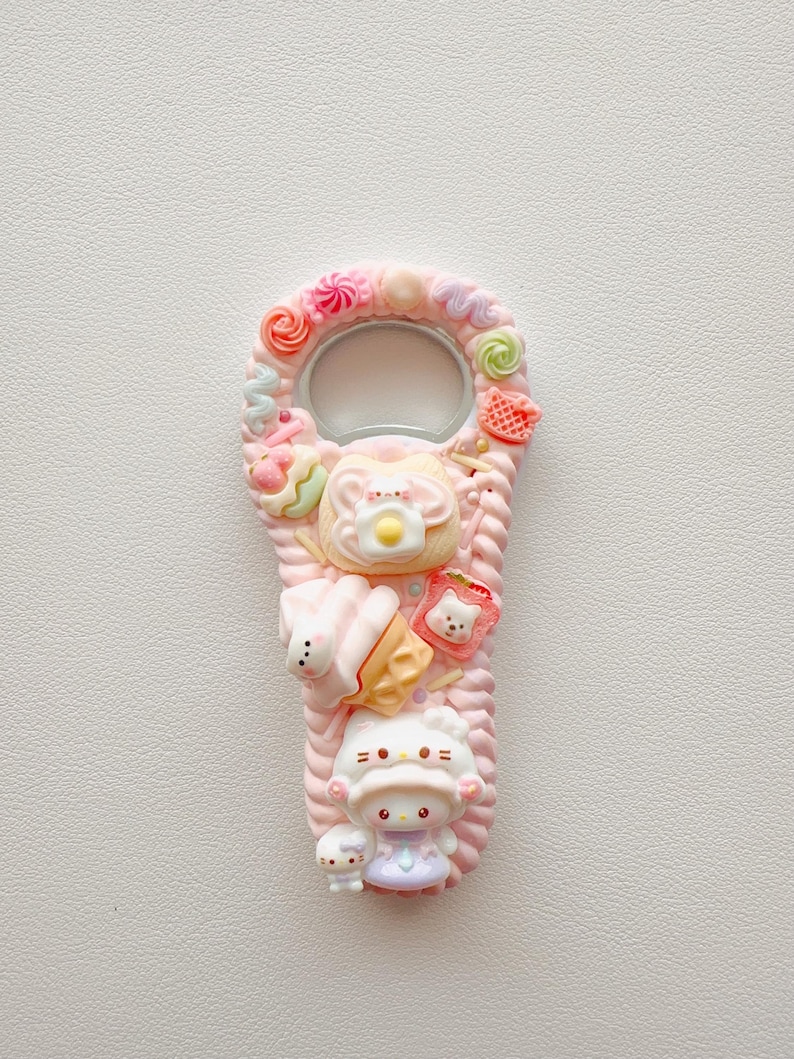 Pu&ograve; includere: Un apribottiglie rosa decorato con ciondoli in resina in miniatura, tra cui una figura di personaggio bianco, un cono gelato, una fetta di pane tostato con un uovo al tegamino e vari vortici colorati e piccole forme di caramelle sui bordi.