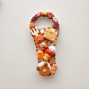 Pu&ograve; includere: Un apribottiglie in metallo decorato con ciondoli in miniatura a tema alimentare, tra cui un orsacchiotto marrone, un cartone di latte con l'etichetta MOM, un piccolo cono gelato, una forchetta e un personaggio con faccia d'orso su base marrone.