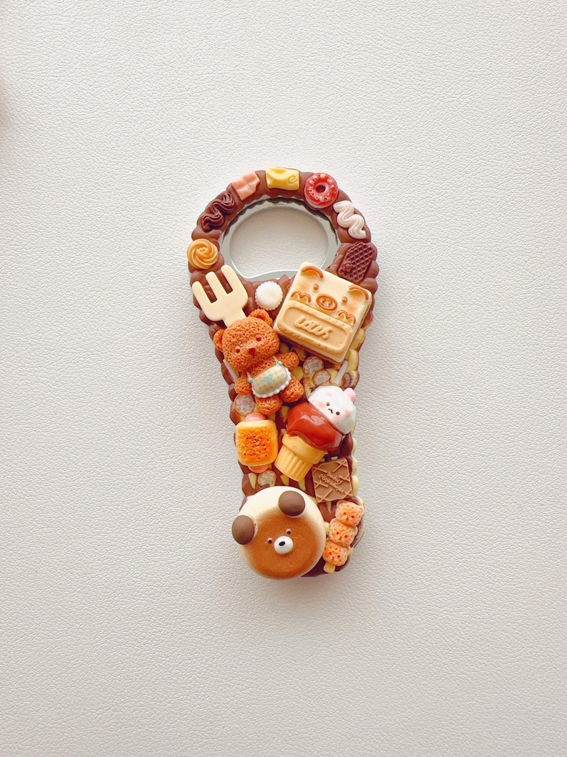 Pu&ograve; includere: Un apribottiglie in metallo decorato con ciondoli in miniatura a tema alimentare, tra cui un orsacchiotto marrone, un cartone di latte con l'etichetta MOM, un piccolo cono gelato, una forchetta e un personaggio con faccia d'orso su base marrone.