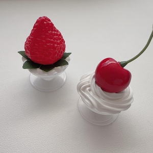 May include: Two decorative ornaments shaped like fruit desserts. One features a bright red textured strawberry resting on green leaves in a white pedestal dish. The other is a glossy red cherry with a green stem atop a swirl of white cream.