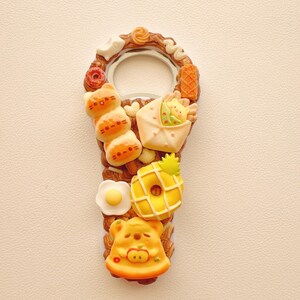 Pu&ograve; includere: Un apribottiglie in metallo decorato con ciondoli in miniatura a tema alimentare, tra cui uno spiedino a tre parti a forma di gatto, un panino incartato, un uovo al tegamino, una ciambella con motivo ad ananas e un pezzo di pizza a forma di orso nei toni del marrone, giallo e bianco.