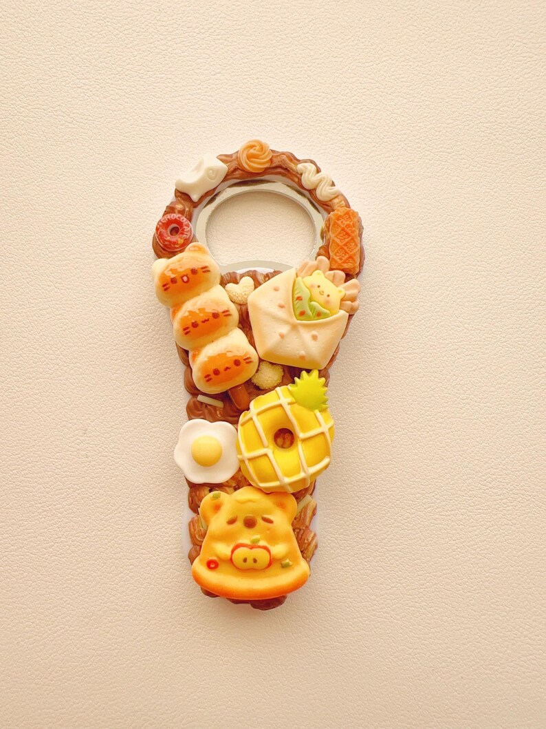Pu&ograve; includere: Un apribottiglie in metallo decorato con ciondoli in miniatura a tema alimentare, tra cui uno spiedino a tre parti a forma di gatto, un panino incartato, un uovo al tegamino, una ciambella con motivo ad ananas e un pezzo di pizza a forma di orso nei toni del marrone, giallo e bianco.
