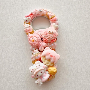 Pu&ograve; includere: Un apribottiglie decorato con varie figurine in miniatura di colore rosa e pastello, tra cui un personaggio gatto, un maiale, un pesce e una creatura soffice. La superficie &egrave; impreziosita da piccole perline e ciondoli nei toni del rosa, bianco e giallo.