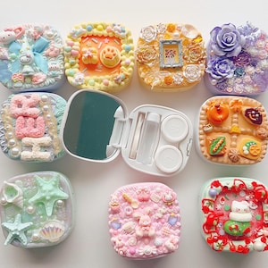 Peut inclure: Une collection de huit étuis à lentilles carrés ornés d'embellissements texturés en 3D aux thèmes variés, incluant des motifs floraux, alimentaires et des personnages, avec un étui ouvert révélant un miroir et des compartiments.