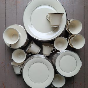 May include: A collection of ivory-colored dinnerware with a silver trim. The set includes plates, saucers, and teacups. The arrangement is on a gray wooden surface, showcasing the elegant design of the tableware.