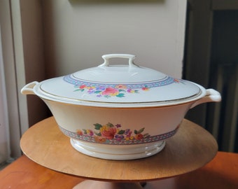 May include: A white ceramic serving bowl with a floral design in shades of pink, purple, and yellow. The bowl has a gold trim and a lid with a handle. The bowl is sitting on a wooden pedestal.