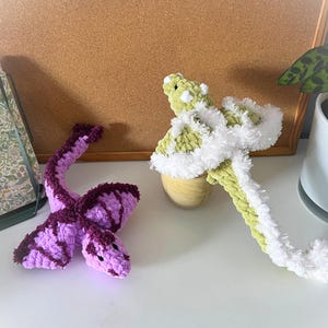 Puede incluir: Dos juguetes de serpiente marina hechos a mano con ganchillo. Uno es morado y burdeos, el otro es verde y blanco. Los juguetes están sobre una superficie blanca con un corcho y un diario decorativo en el fondo.
