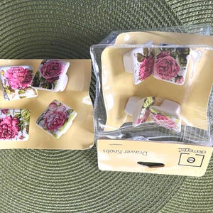 May include: A set of eight square drawer knobs featuring a pink rose floral pattern and vintage-style script text. Four knobs are displayed on a yellow cardboard backing, and four are enclosed in a clear plastic package labeled Drawer Knobs.
