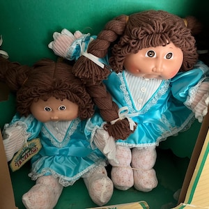 May include: Two Cabbage Patch Kids dolls with brown yarn hair styled in pigtails, wearing matching bright blue satin dresses with white lace trim and white patterned tights. The dolls are positioned inside a green and yellow cardboard display box.