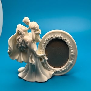May include: Ivory-colored photo frame featuring a mother figure holding a child, with an oval frame decorated with floral accents. The sculpture is set against a blue background, creating a serene and sentimental display.