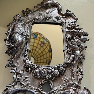 May include: Ornate silver-toned vanity mirror with a rectangular glass. The frame features sculpted figures, floral designs, and cherubs. The mirror has a vintage, antique aesthetic.