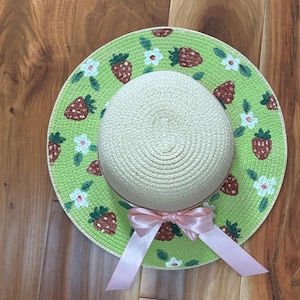 May include: A straw sun hat with a light green brim decorated with red strawberries and white flowers. The hat has a beige crown and a pink ribbon bow. The hat is designed for sun protection and adds a touch of style.