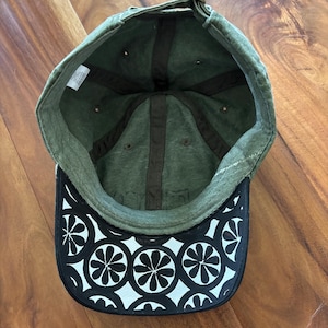 Könnte beinhalten: Grüne Baseballkappe mit einem schwarz-weißen Blumenmuster auf der Unterseite des Schirms. Die Kappe hat einen gebogenen Schirm und einen verstellbaren Riemen am Rücken. Die Innenseite ist mit einem dunkelbraunen Stoff gefüttert.