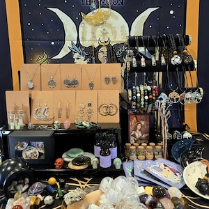 May include: A display of various handcrafted jewelry and crystals. Necklaces, earrings, and bracelets are arranged on stands. A variety of crystals and stones are displayed on a tray. The backdrop features a celestial tapestry with the text "Base Items" and "The Moon".