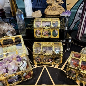 May include: Several ornate, gold-colored treasure chests filled with colorful crystals and gemstones. The chests are displayed on a black surface with a gold geometric pattern, alongside other crystals and decorative items.