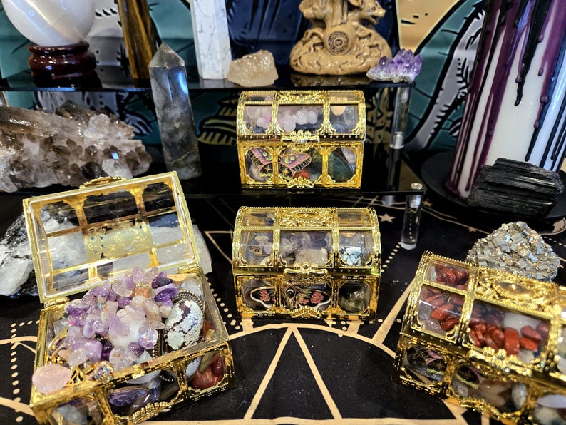 May include: Several ornate, gold-colored treasure chests filled with colorful crystals and gemstones. The chests are displayed on a black surface with a gold geometric pattern, alongside other crystals and decorative items.