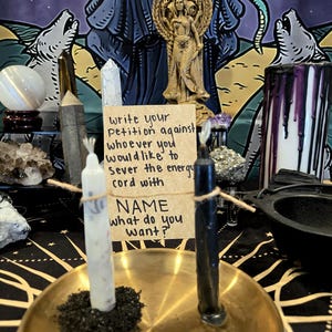 May include: A spiritual ritual setup featuring a gold-colored brass tray holding a white and a black candle. The candles are connected by twine to a handwritten note that reads, "Write your petition against whoever you would like to sever the energy cord with NAME what do you want?"