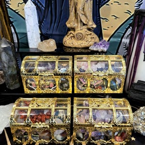 May include: Four small, ornate gold treasure chests with clear tops, each filled with colorful gemstones. The chests are arranged on a black surface, with a statue and crystals in the background. The chests have decorative gold accents.